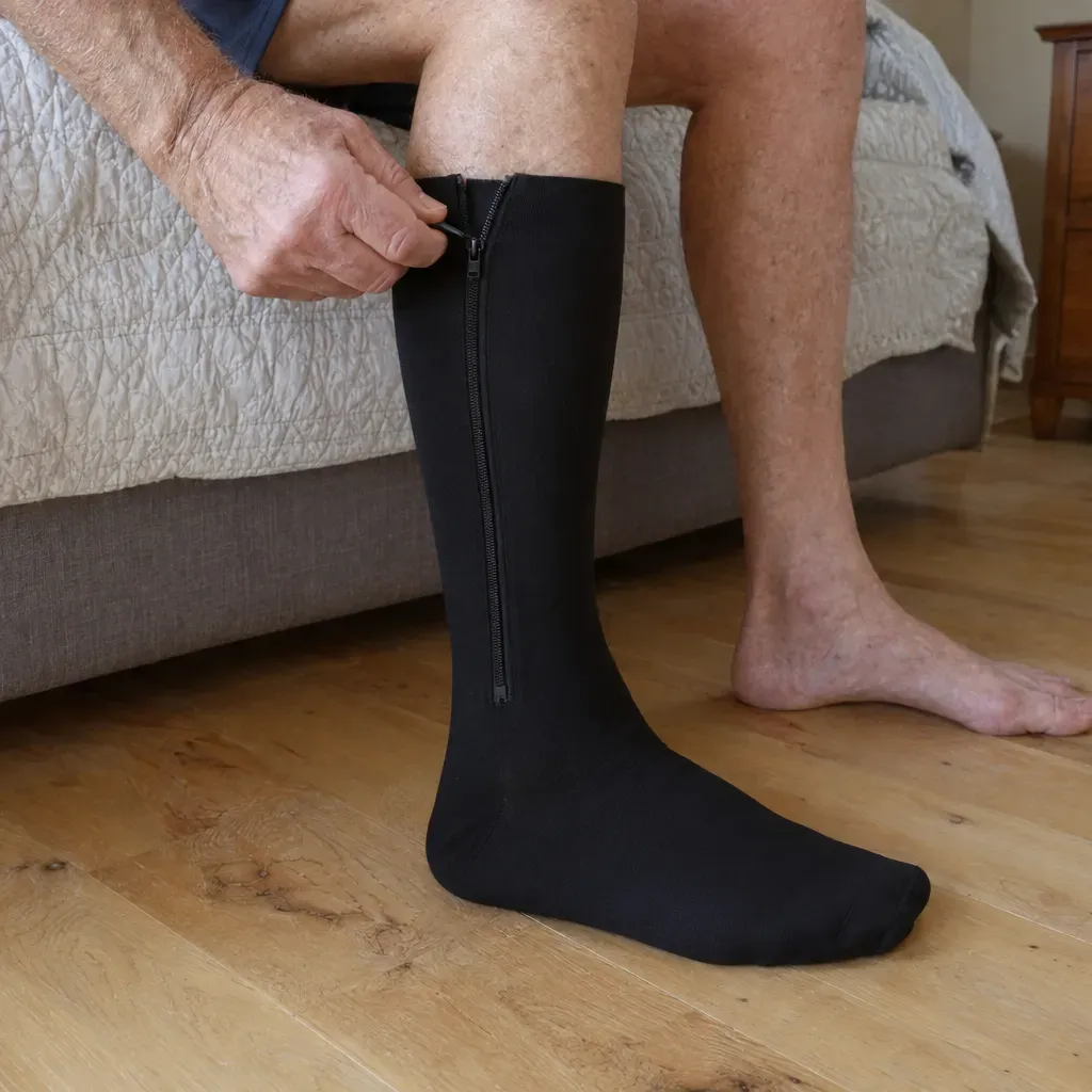 An older man seated on his bed zipping up a black zipper compression sock